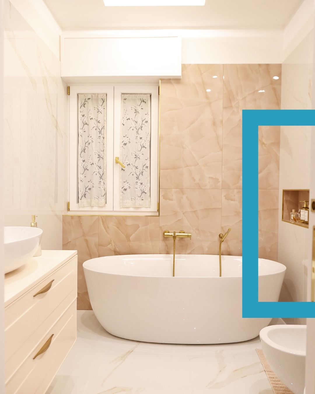 Bagno luminoso color beige con vasca freestanding bianca, finestra, mobile lavabo e nicchia doccia con dettagli blu.