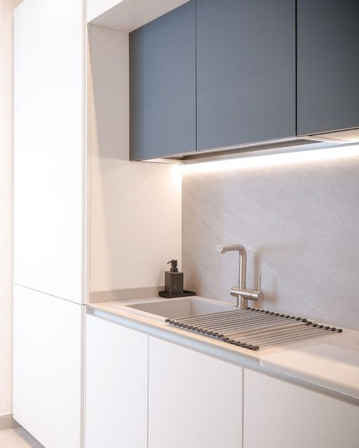 Cucina moderna e minimalista con mobili bianchi, pensili scuri, lavello e illuminazione sottopensile.