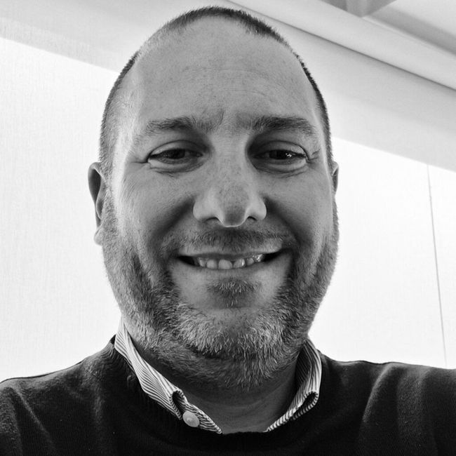Uomo sorridente con camicia e maglione, ritratto in un selfie in bianco e nero in primo piano.