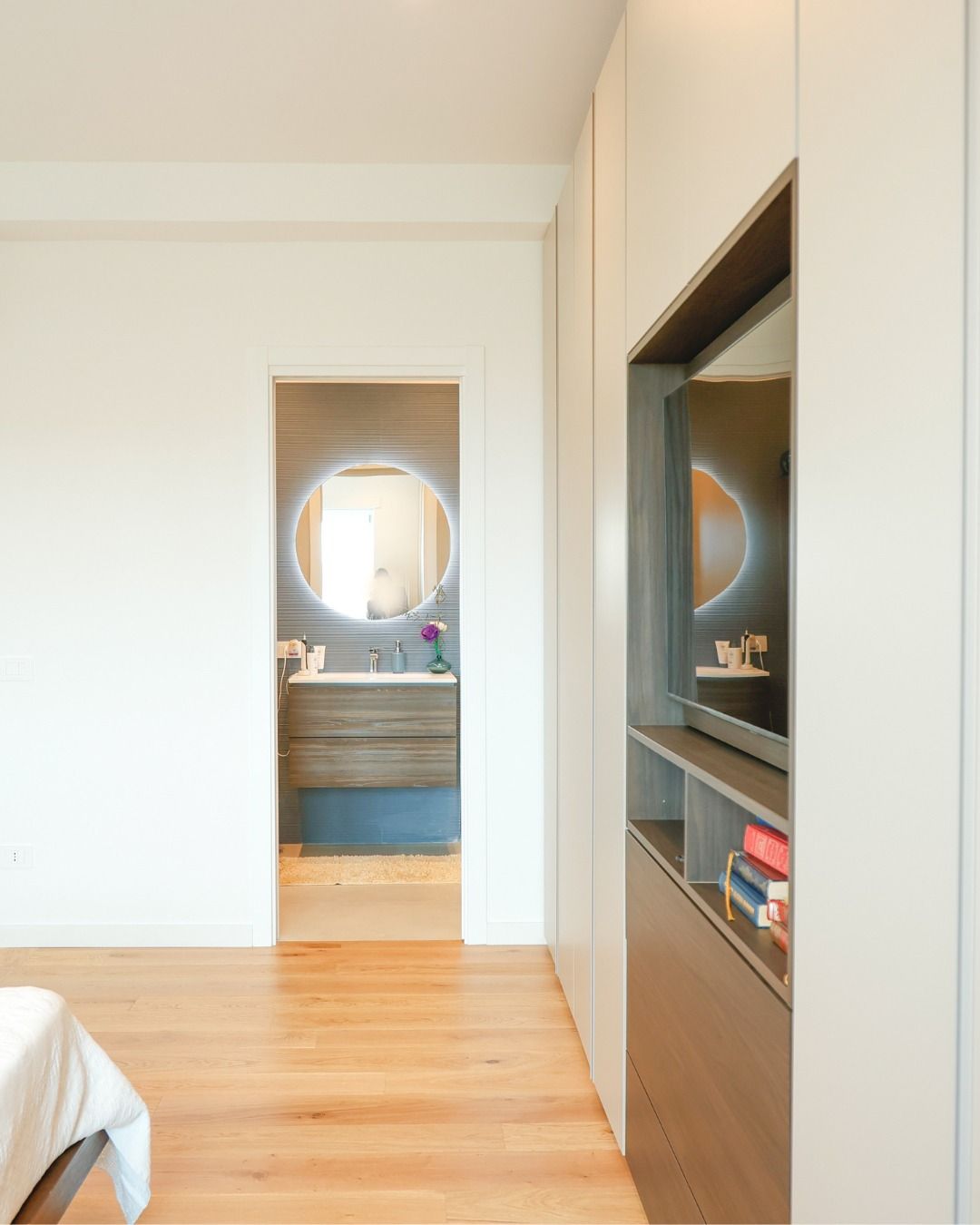 Luminosa camera da letto moderna con pareti bianche, pavimento in legno e porta che conduce al bagno con specchio rotondo.