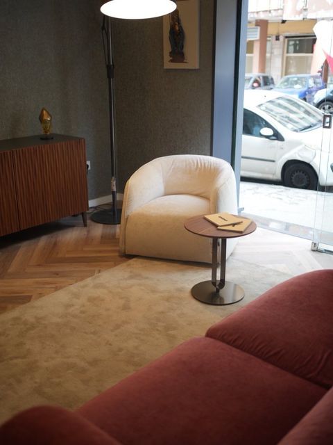 Accogliente salotto con divano rosso, poltrona bianca, tavolino rotondo, lampada e grande finestra con vista sulle auto parcheggiate.