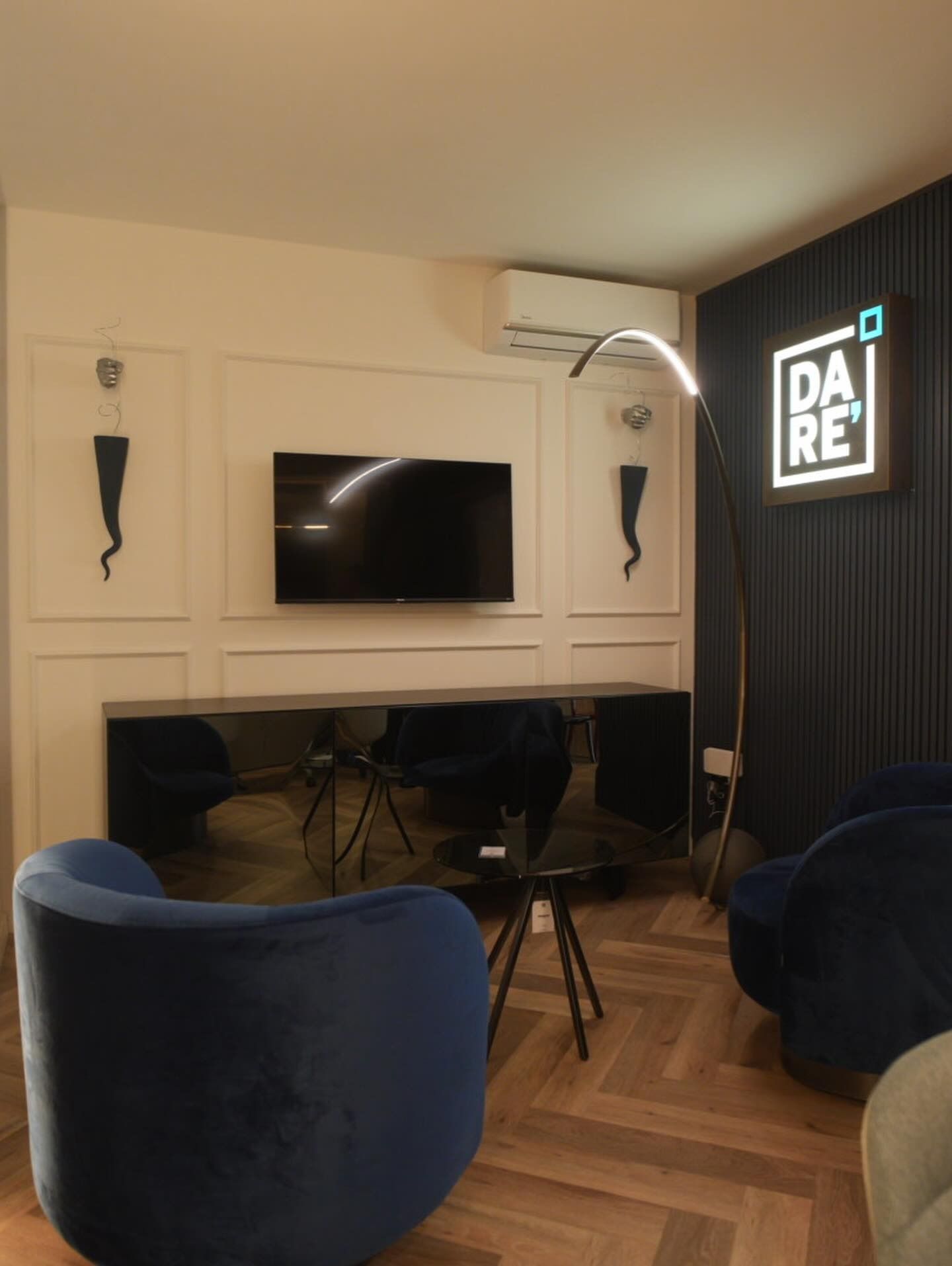 Salotto moderno con TV a parete, tende scure, sedie blu e un'insegna luminosa con la scritta 