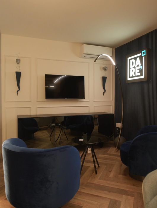 Salotto moderno con TV a parete, tende scure, sedie blu e un'insegna luminosa con la scritta 