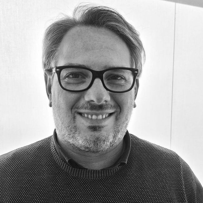 Ritratto in bianco e nero di una persona sorridente con occhiali e maglione a trama grossa