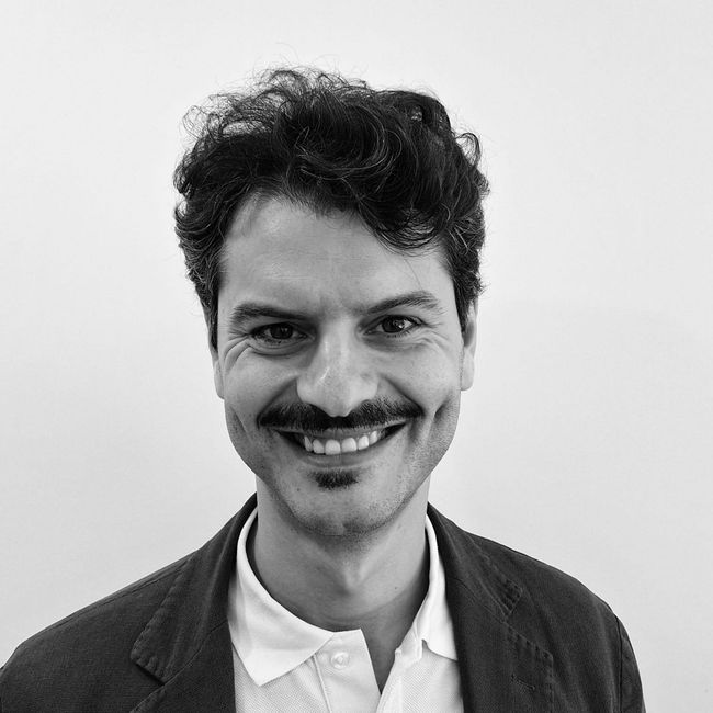 Ritratto in bianco e nero di un uomo sorridente con capelli scuri e baffi, che indossa una camicia con colletto e una giacca.