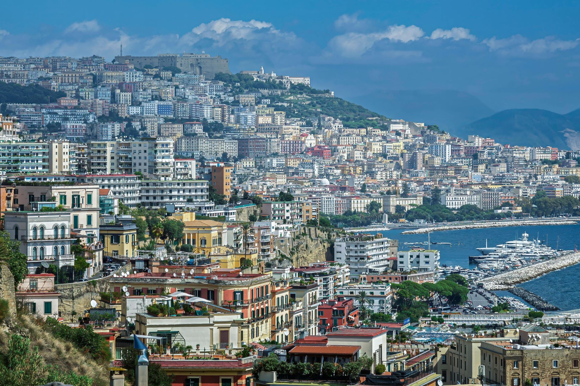 Città costiera panoramica con edifici arroccati sulle colline, barche del porto turistico e montagne sullo sfondo.
