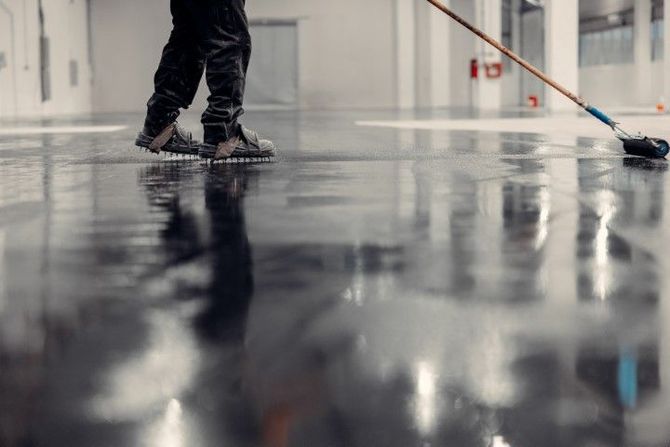 A person wearing spiked shoes uses a roller to apply a glossy, dark coating to a large warehouse floor.