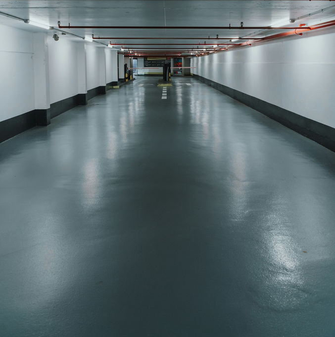 A long, narrow indoor parking garage with smooth, light-gray flooring, white walls with black trim, and overhead lighting.