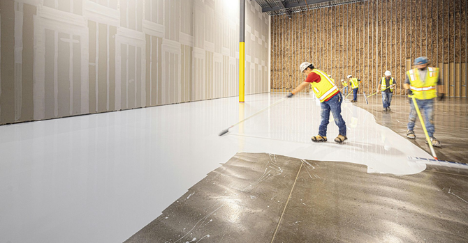 Construction workers in safety gear apply a smooth white epoxy coating to a large concrete warehouse floor.