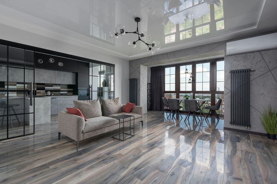 A modern living room with gray wood-look flooring, a beige sofa, dark metal accents, and a dining area by large windows.
