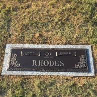 A rectangular, bronze, flat-marker gravestone for the Rhodes family set in grass, featuring names and floral motifs.