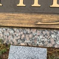 A wooden sign with light-colored letters sits above a speckled granite base, placed near some grass.