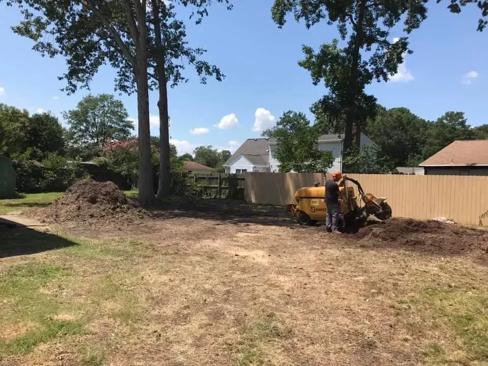 Backyard with dirt clearing, trees, a fence, and a worker operating a yellow excavator