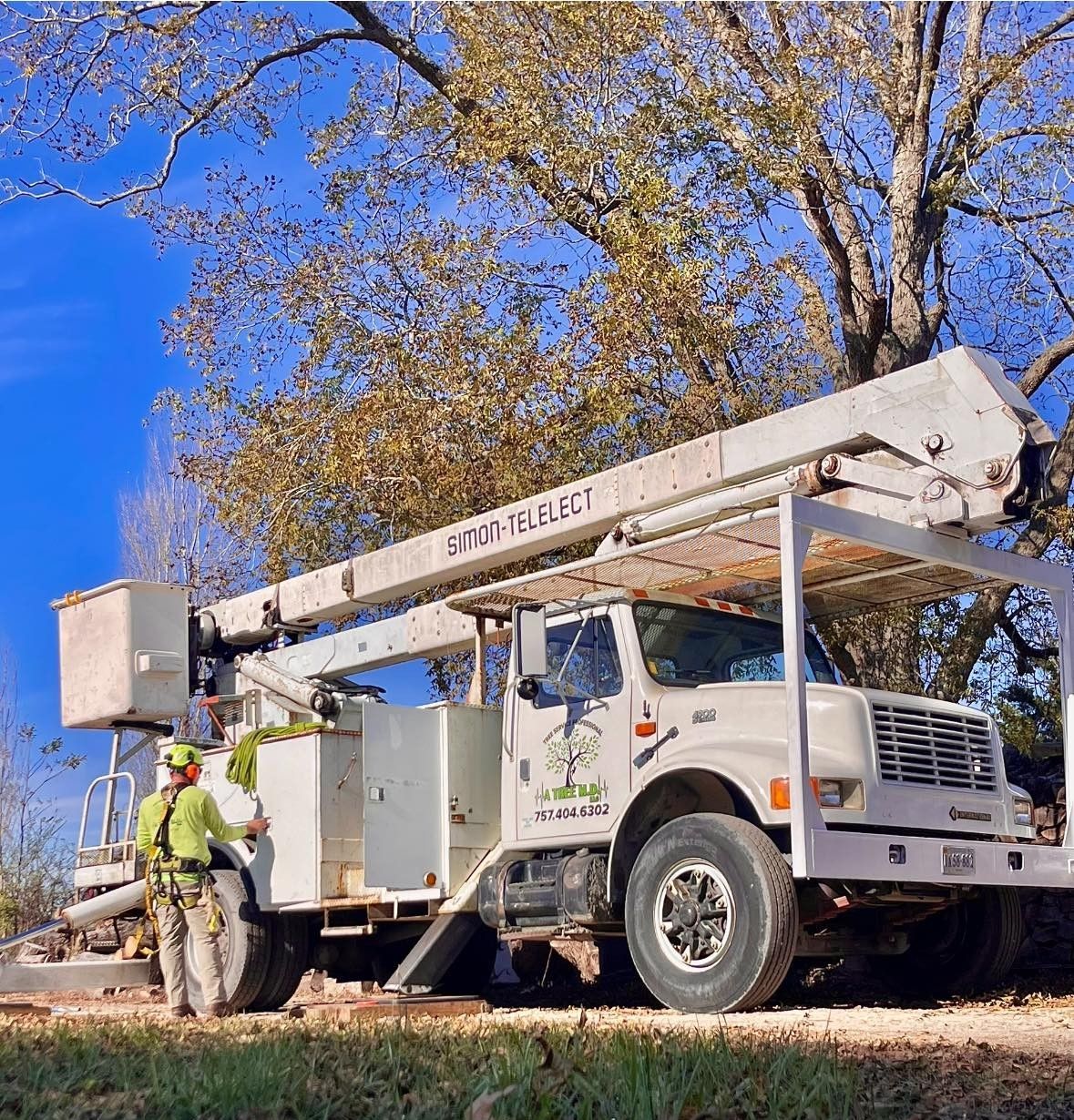 Utility truck with raised bucket arm and workers beside it under a tree on a sunny day