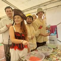 A girl in a pirate hat is smiling and next to her are boys with smiles on their faces mixing something colorful in bowls.