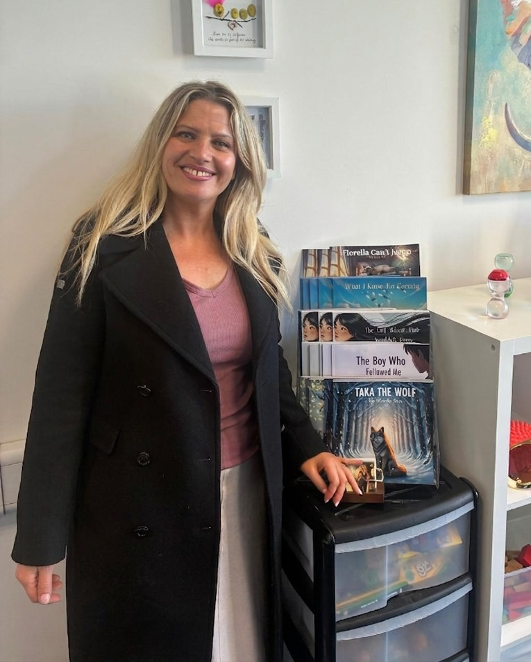 A woman with long blond hair is standing near a shelf with books.