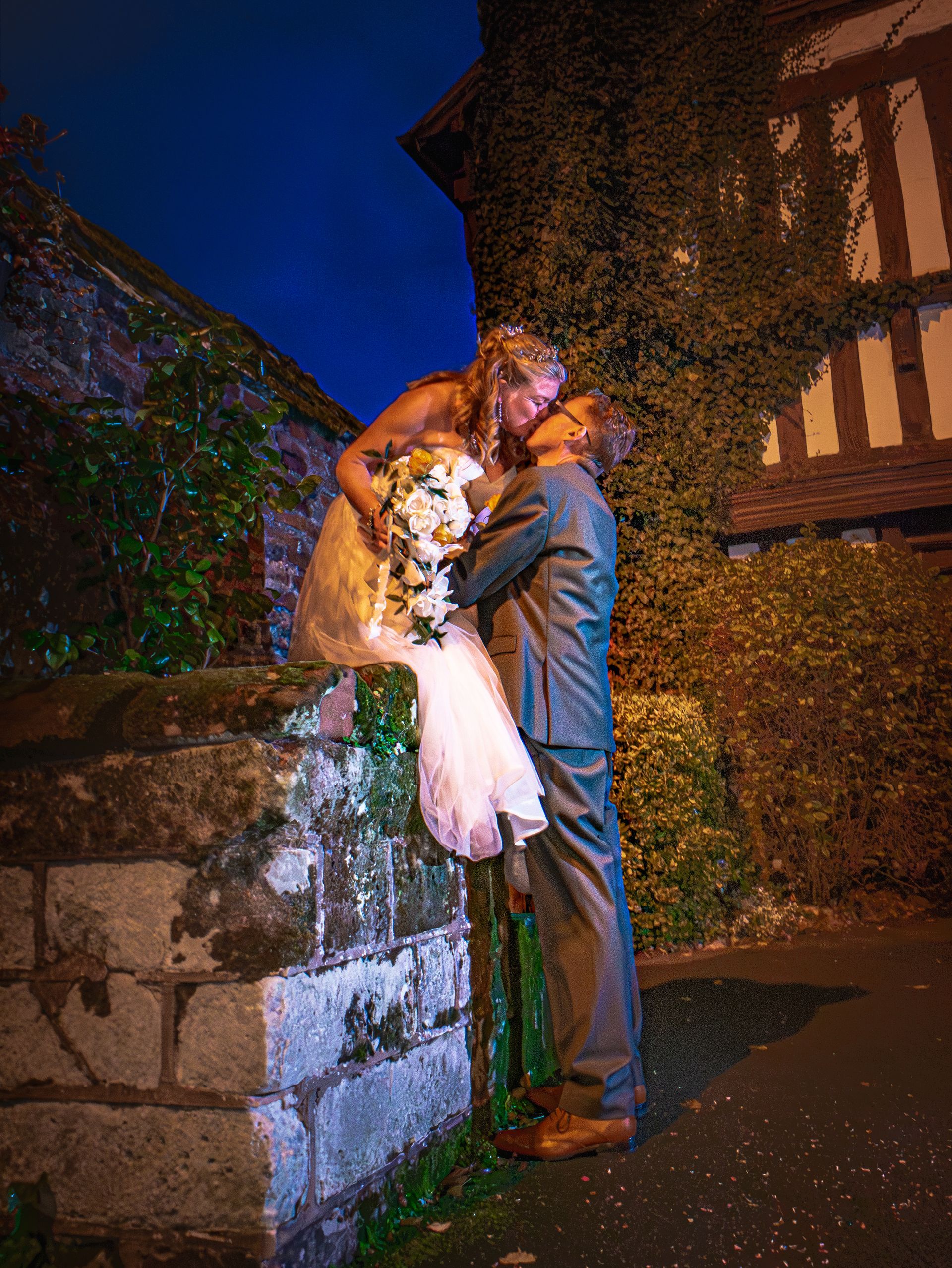 Bride and groom embrace, kissing on a brick wall at night, in front of a building with vines.