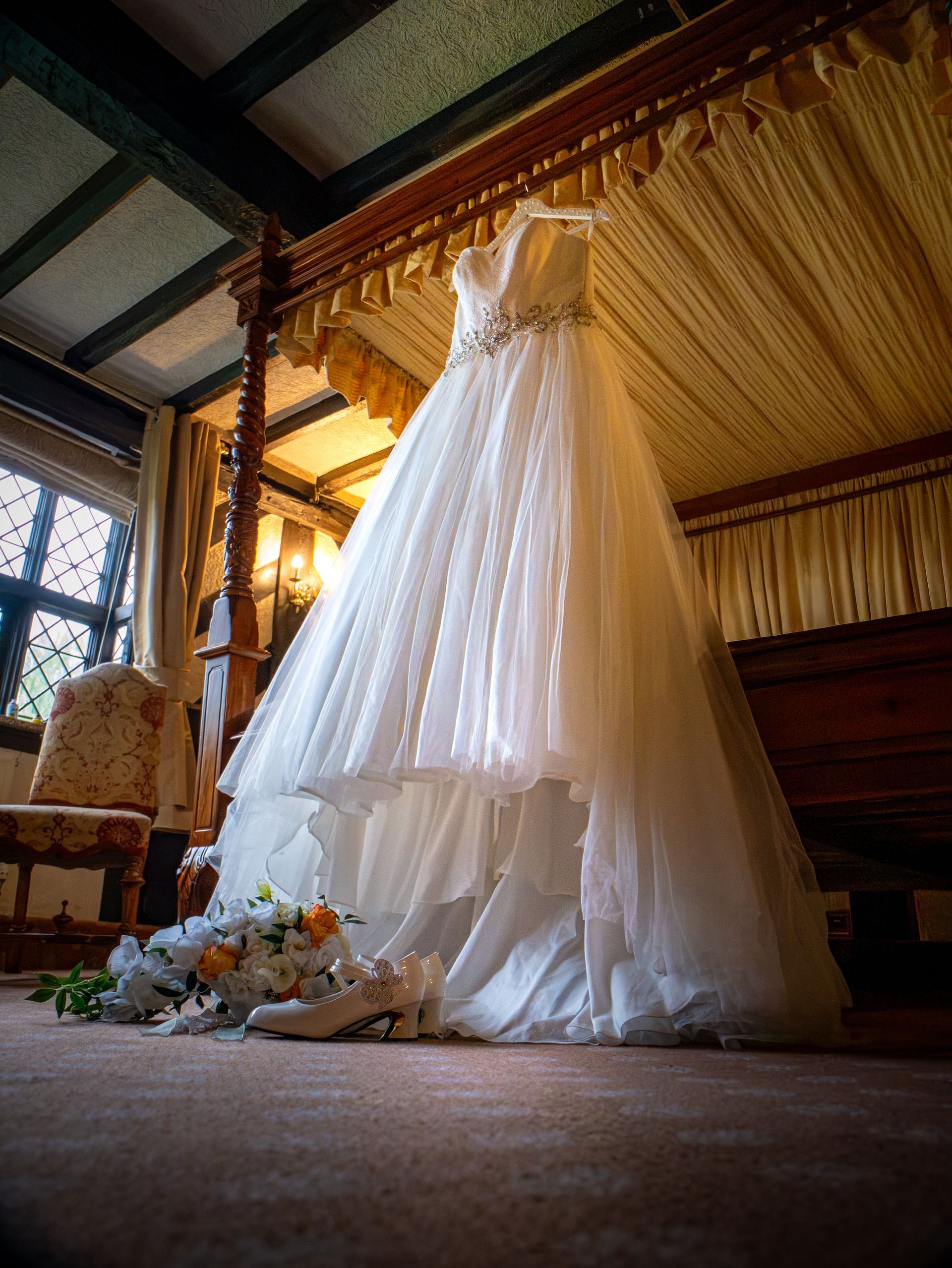 Wedding dress hanging on a wooden four-poster bed, with flowers and shoes at the base.