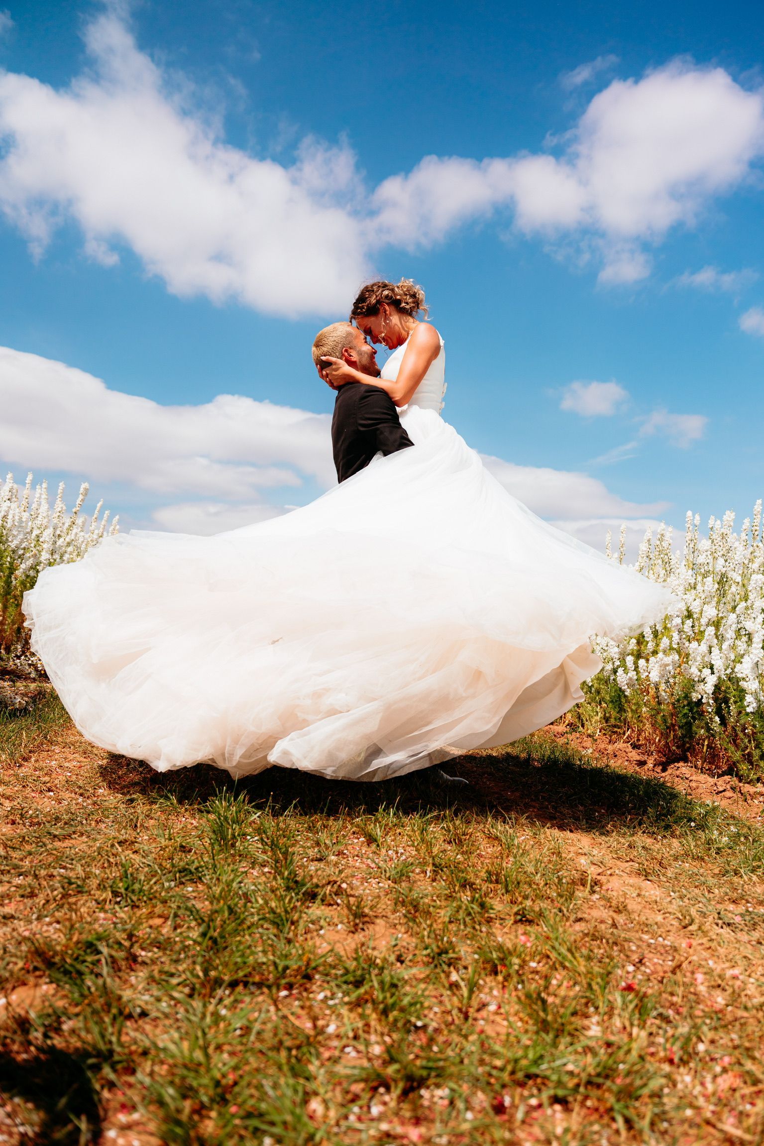 Couple in a field of pink flowers, looking at each other, under a blue sky with clouds.