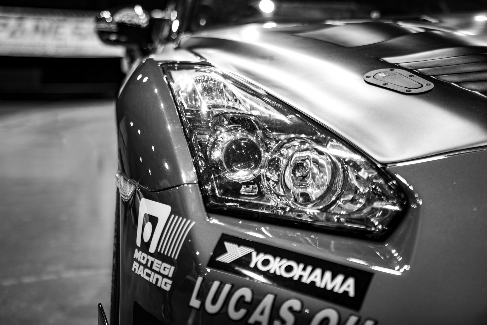 Black and white close-up of a race car's front, showing a headlight and sponsor stickers.