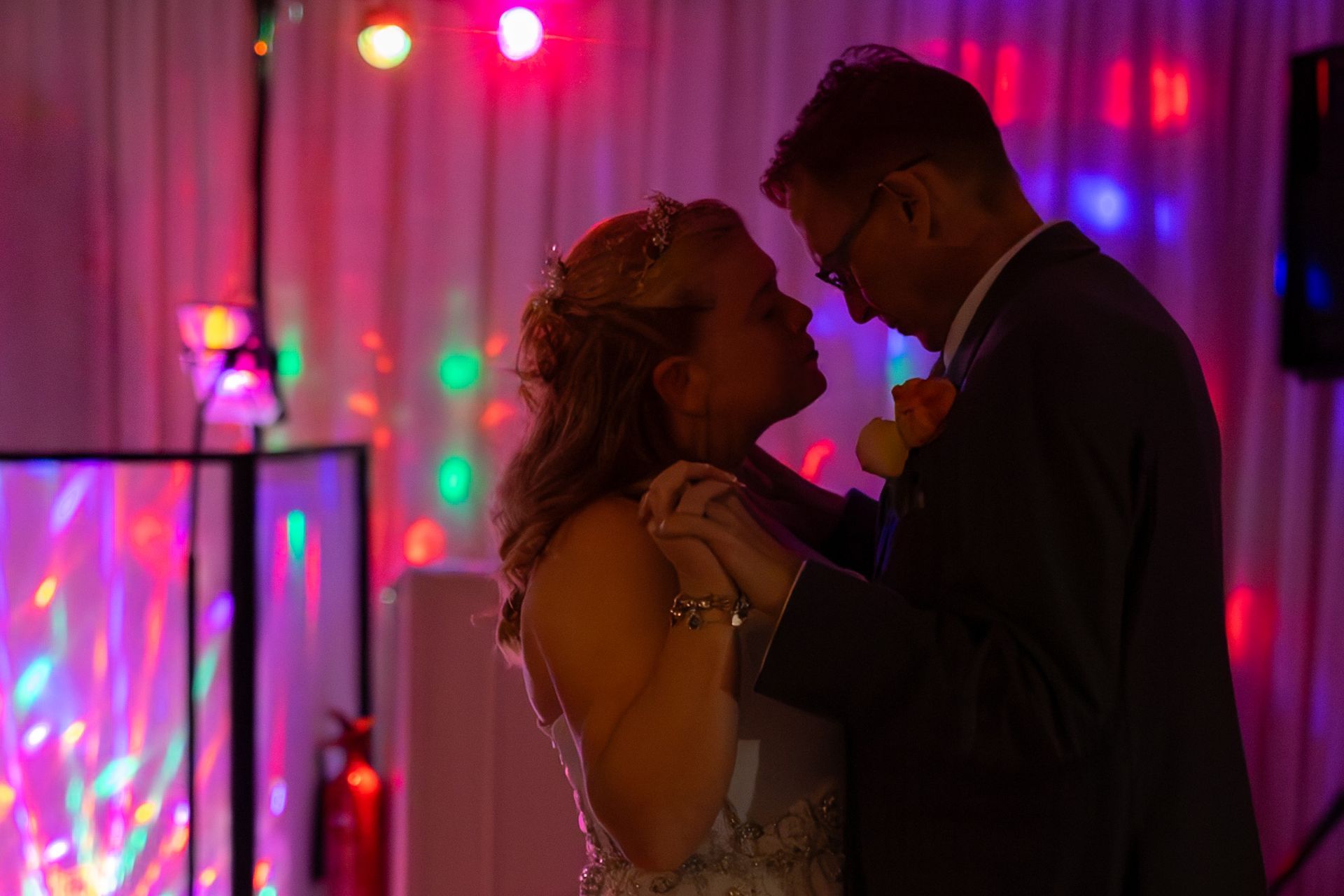 Couple slow dancing at a wedding reception; colorful lights in background.