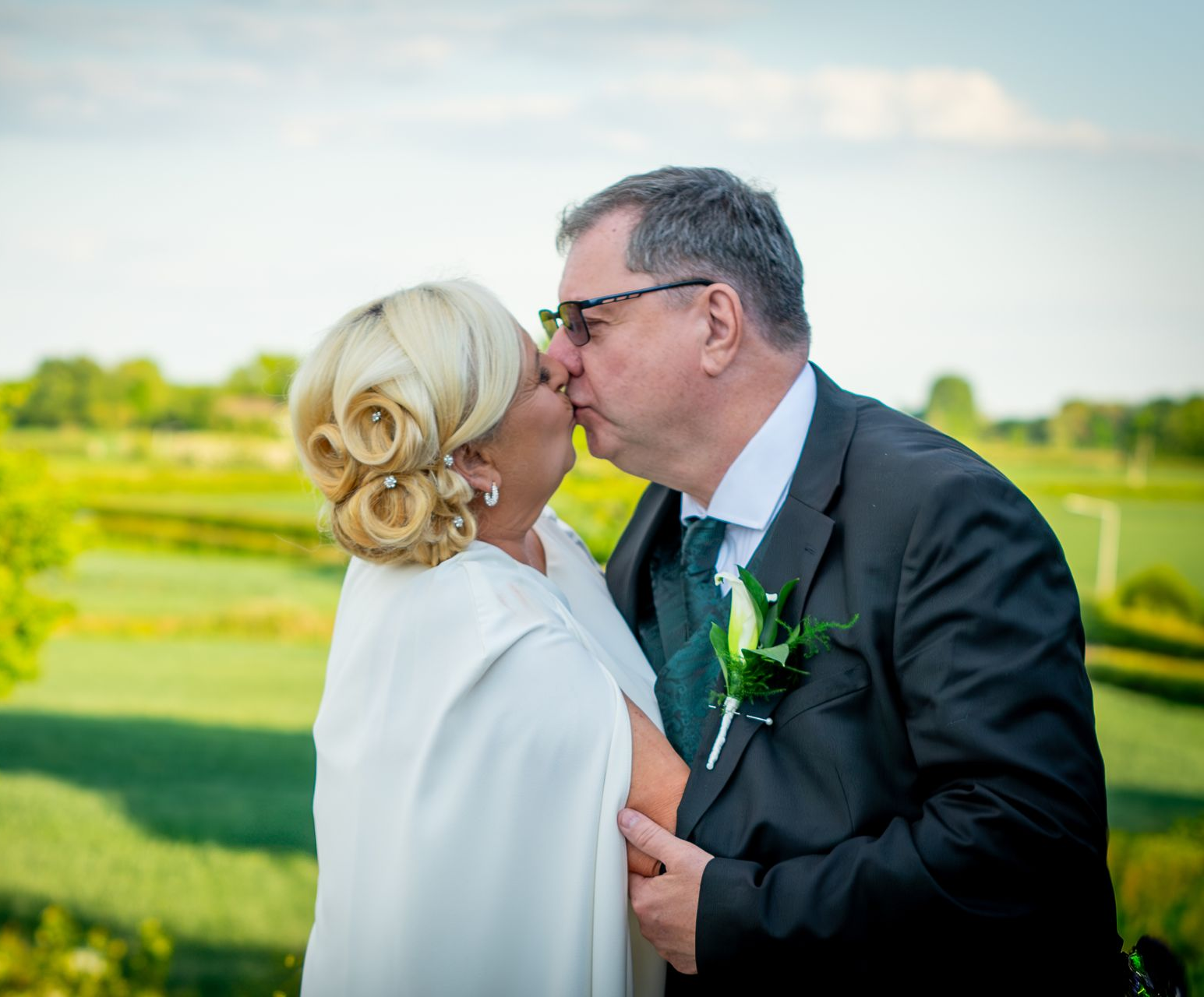 Couple kissing outdoors, bride in white, groom in a suit, green field in the background.