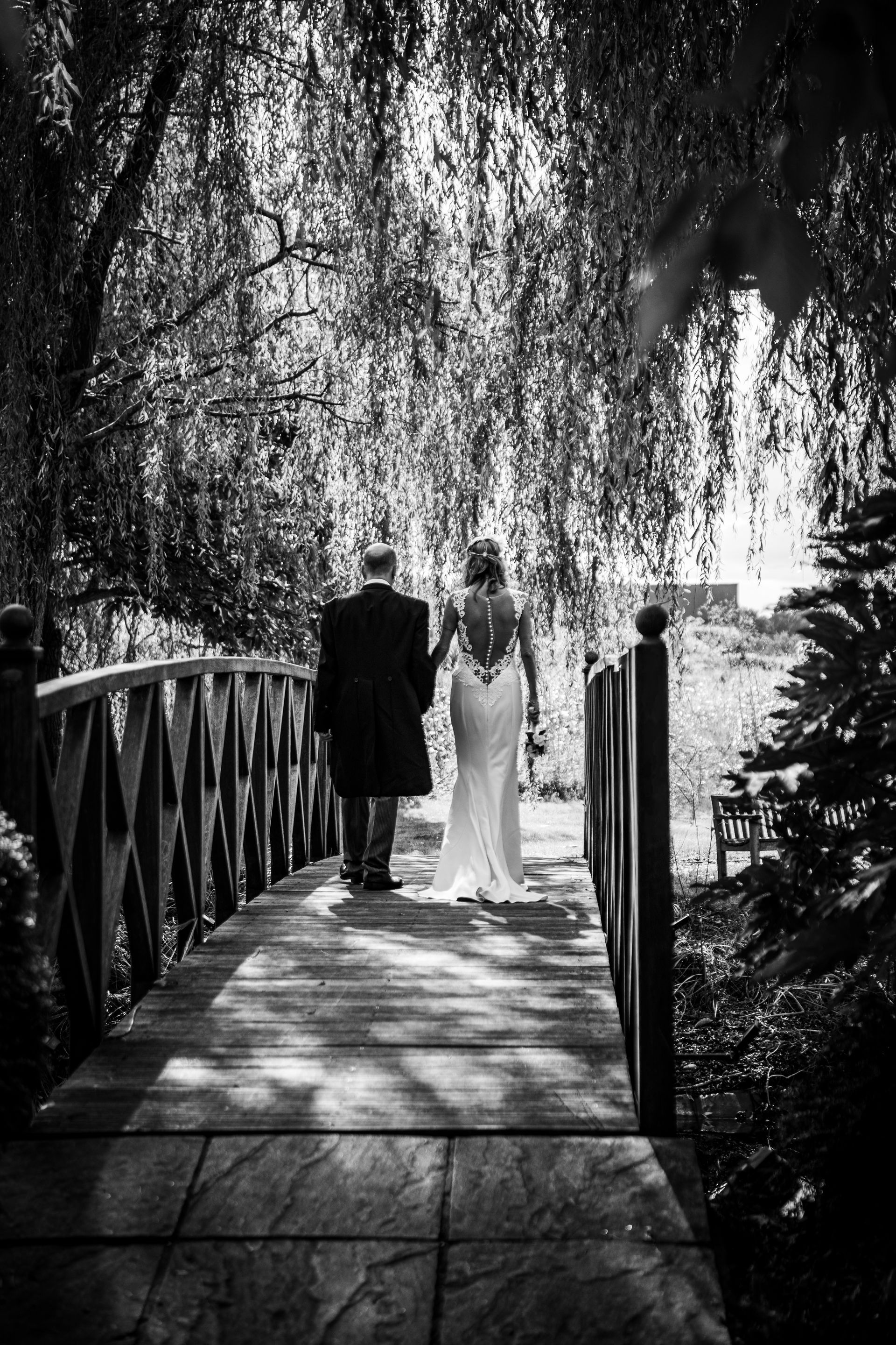 Bride and groom walking on a wooden bridge, back to camera, under weeping willow trees.
