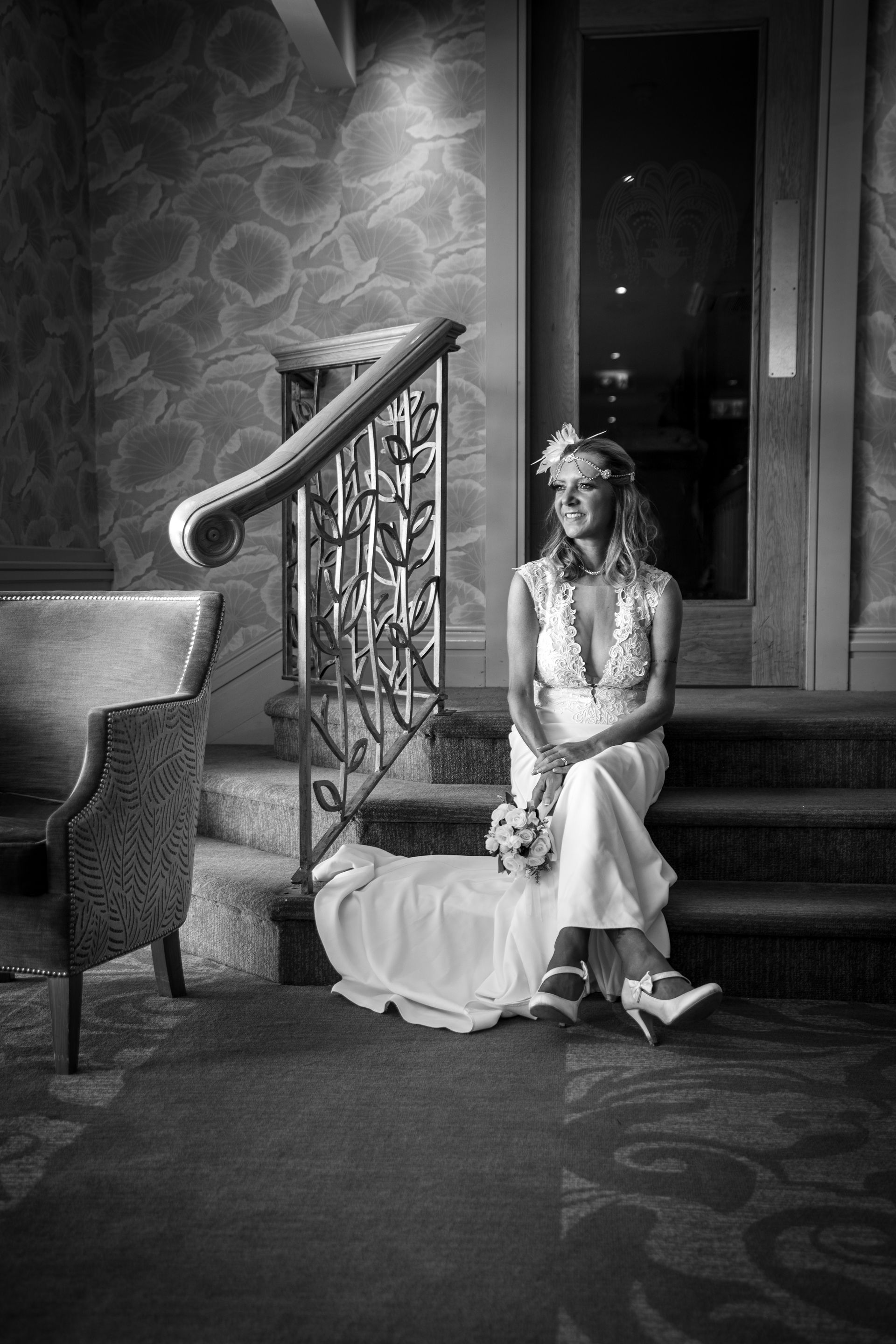 Woman in wedding dress sits on steps, holding bouquet, looking off to the side, near ornate railing. Black and white.