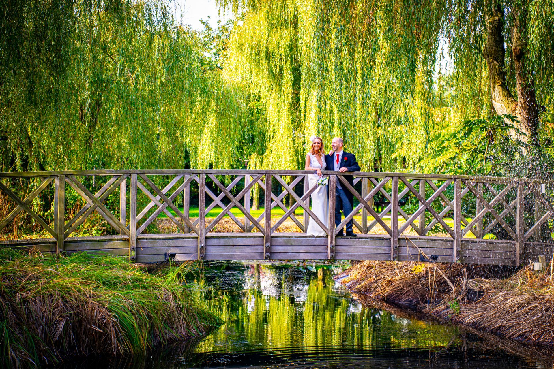 Bride and groom on a wooden bridge, surrounded by greenery and water.