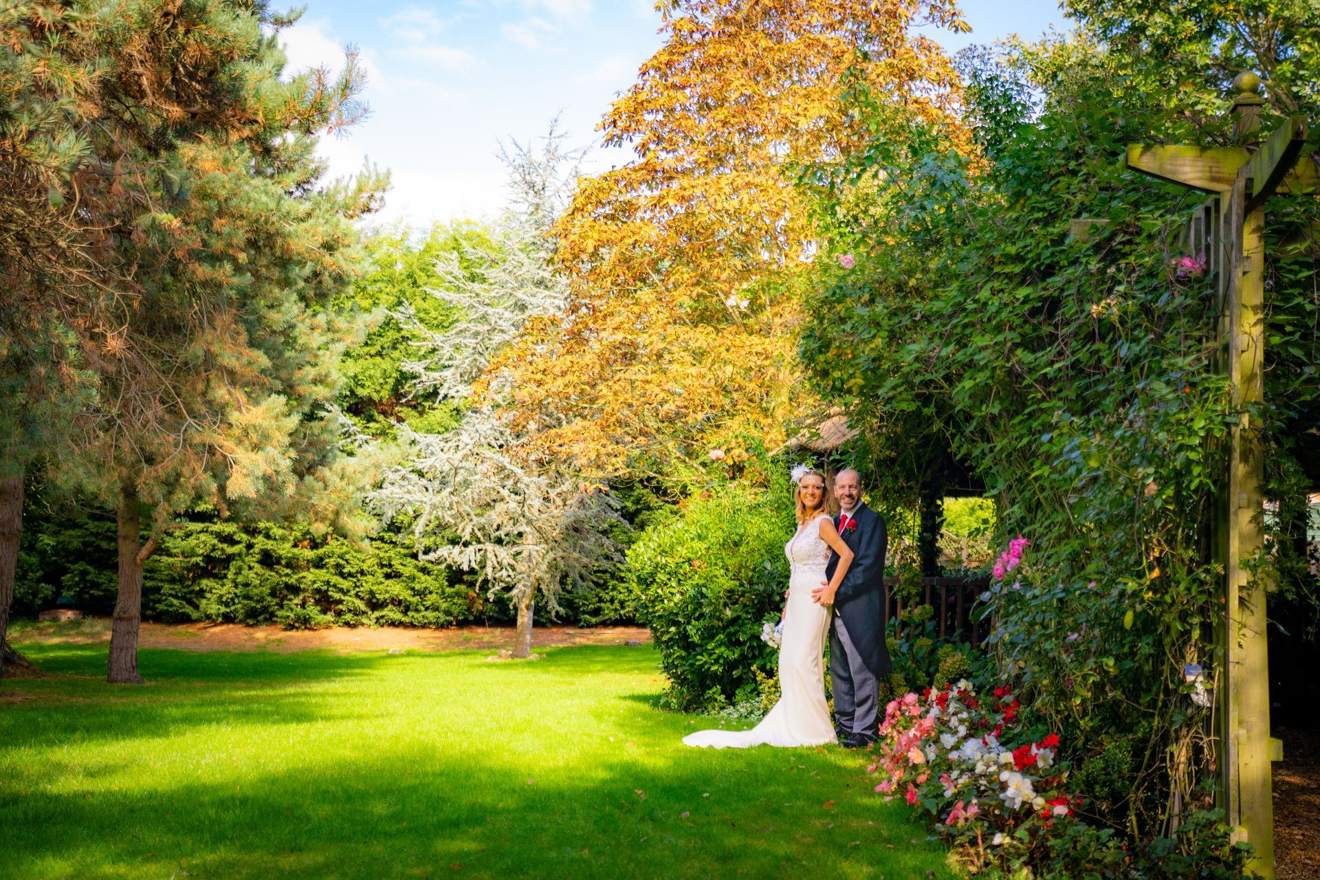 Bride and groom in wedding attire pose in a sunlit garden, surrounded by trees and greenery.