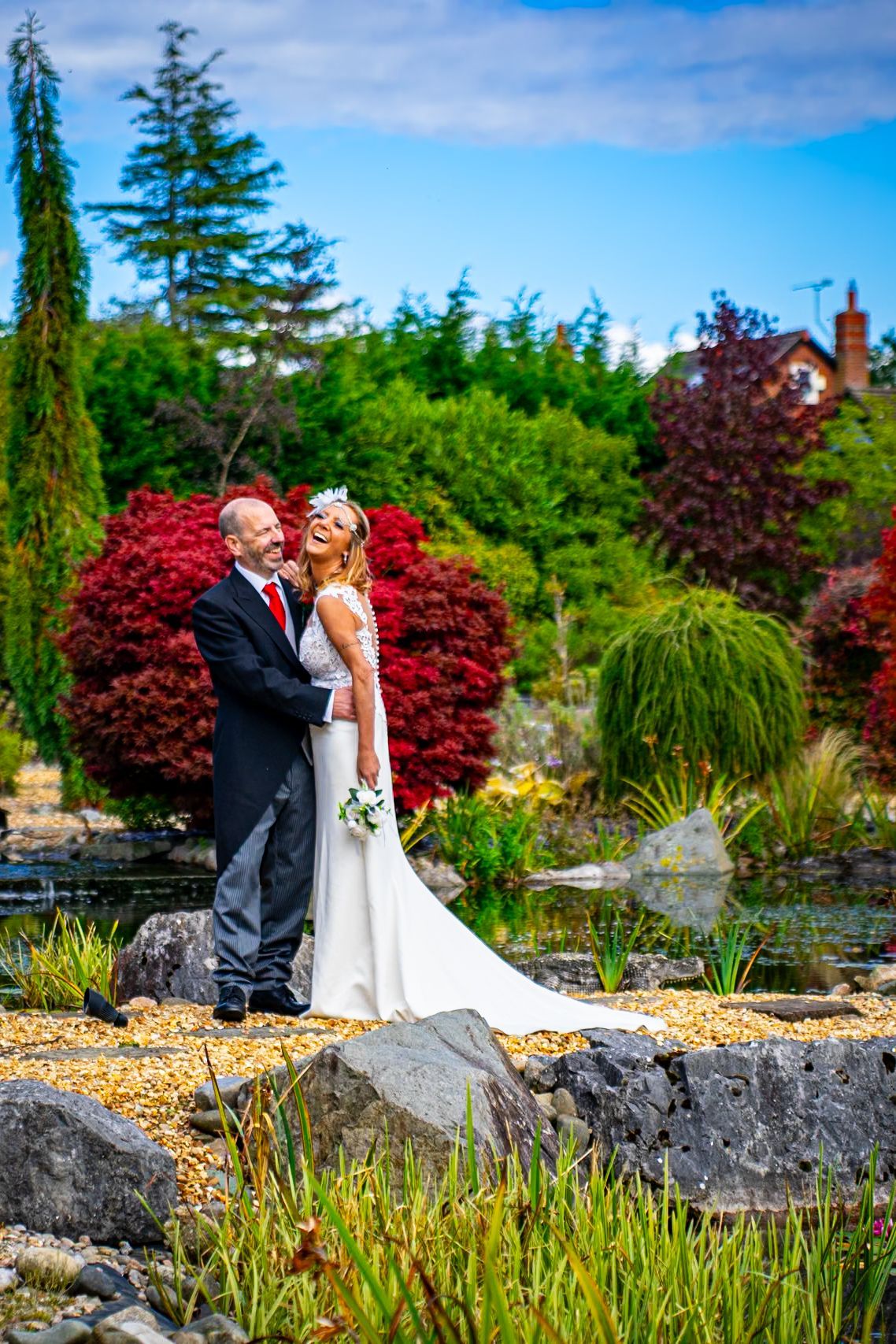 Bride and groom embrace in a garden. Bride laughs, wearing a white dress and flower crown. Groom in black suit, red tie.