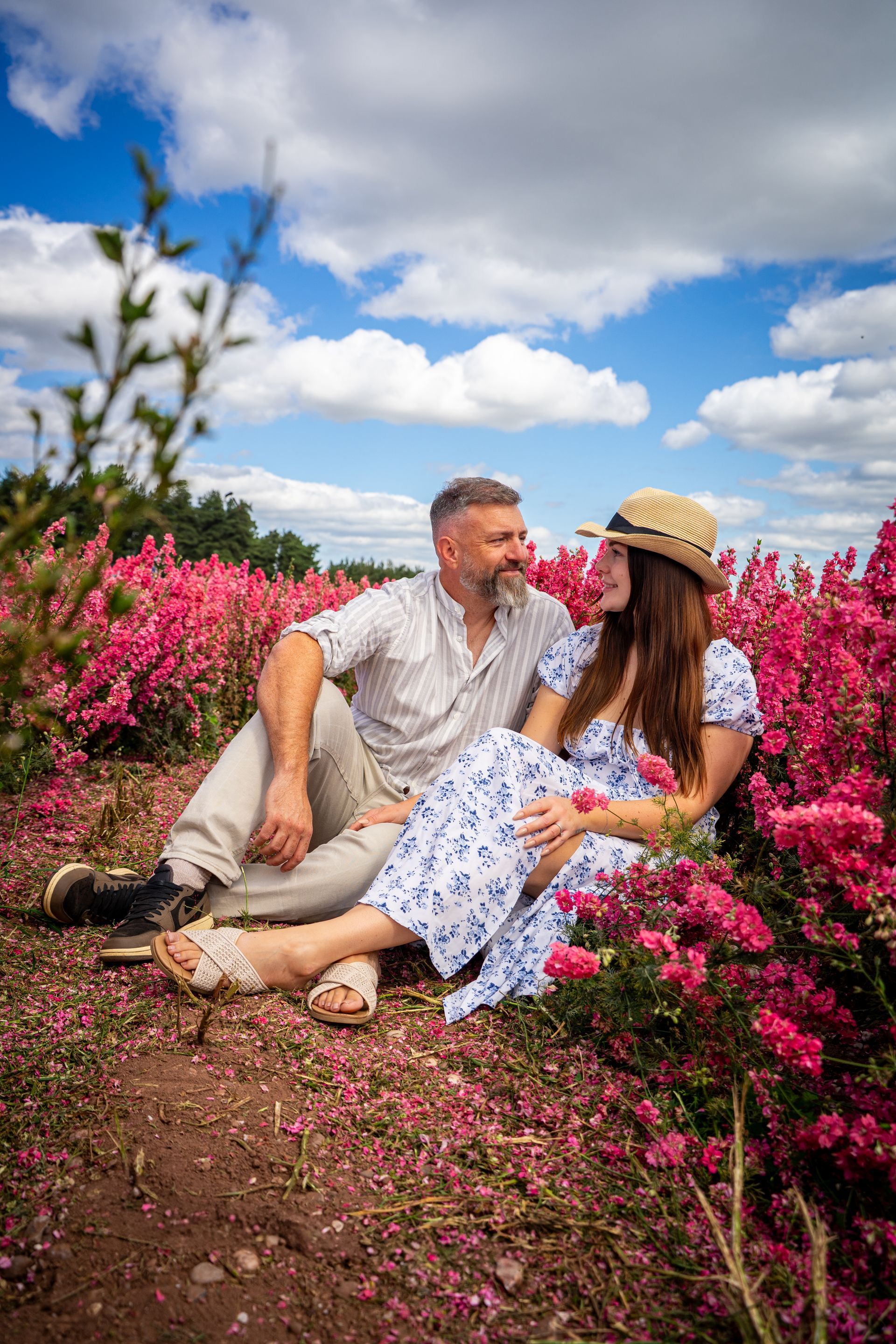 Couple sitting in a field of pink flowers, gazing at each other under a blue, cloudy sky.