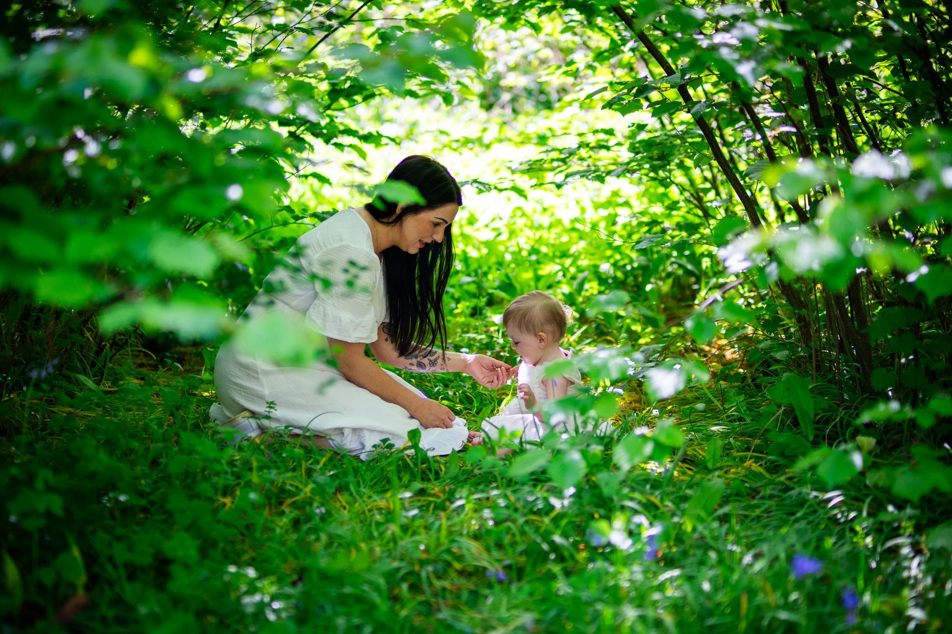 Woman in white dress and baby in grass, surrounded by green foliage.