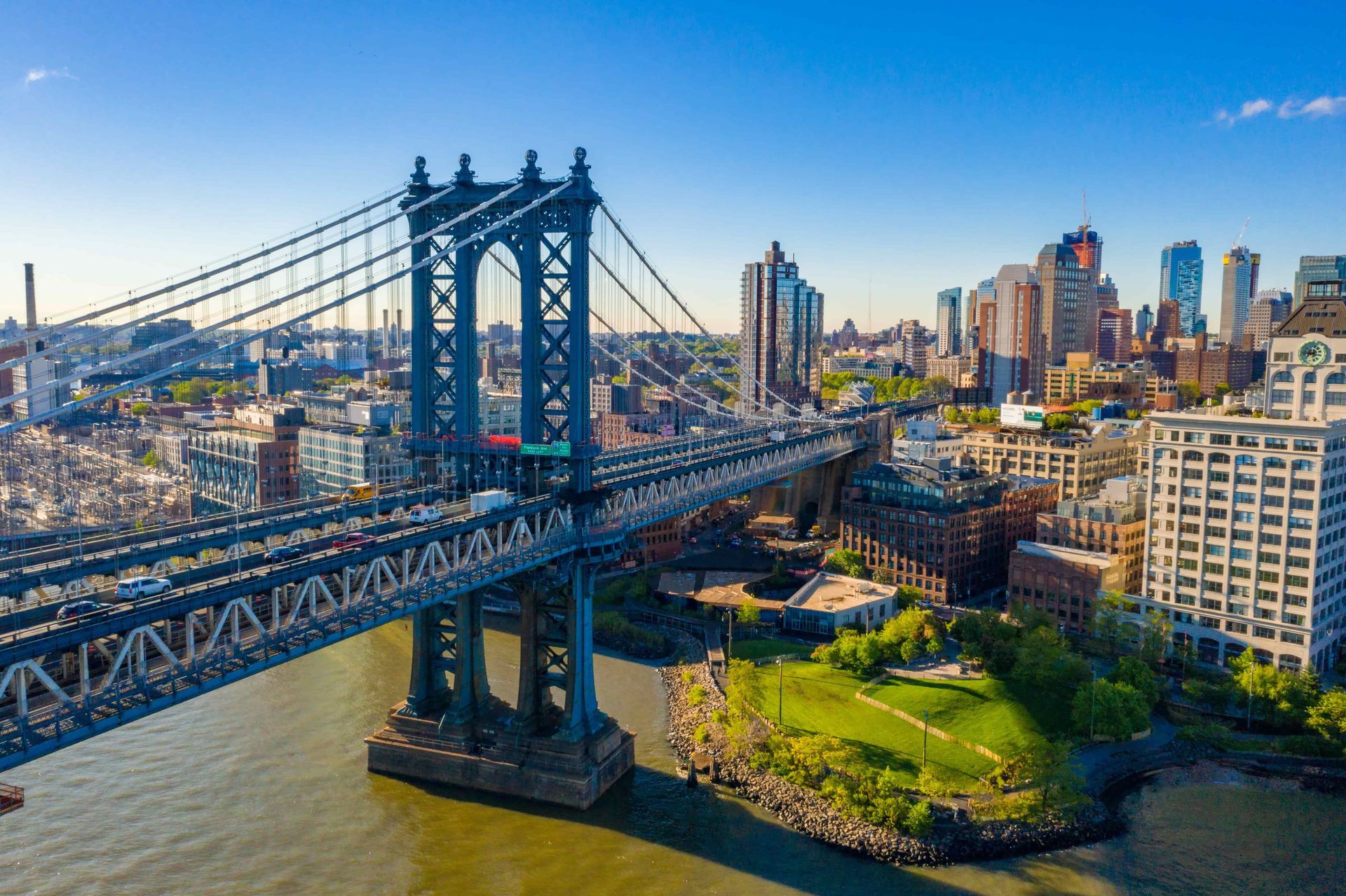 Manhattan Bridge with city skyline in the background, blue sky, and water.