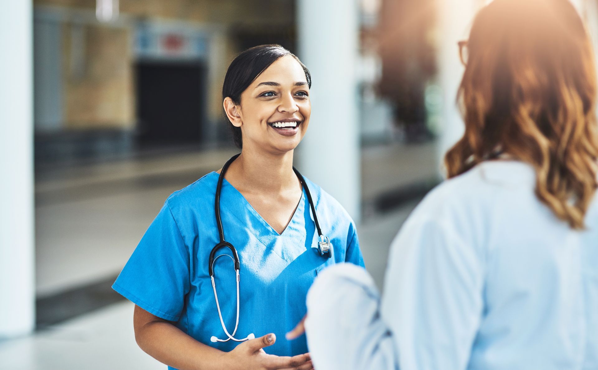 Nurse in blue scrubs smiling while talking to another person in a hospital hallway.