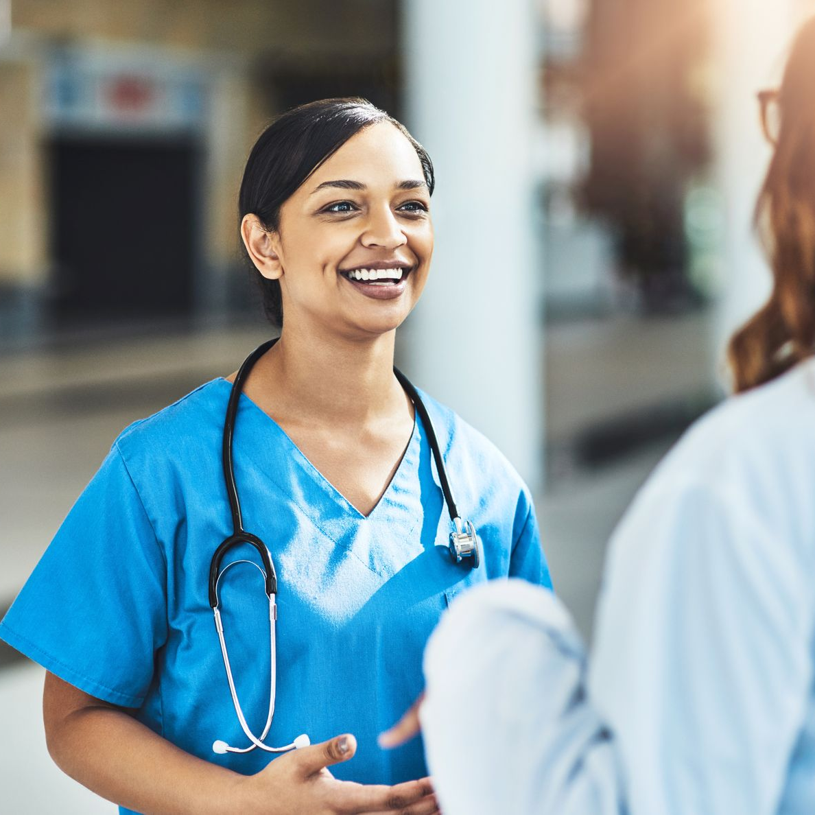 Nurse in blue scrubs with a stethoscope smiles while conversing with another person in a light-colored lab coat.