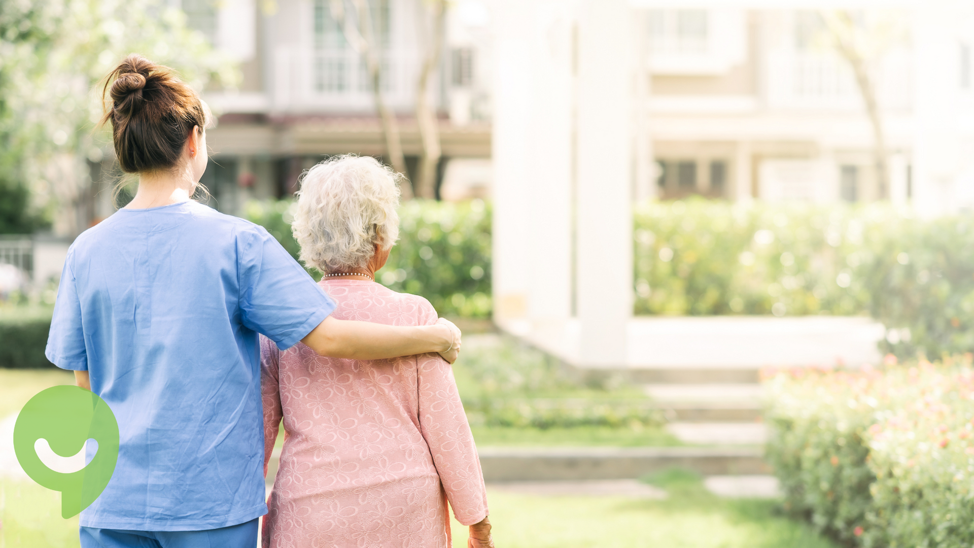 Woman in blue scrubs assists elderly woman walking outdoors.