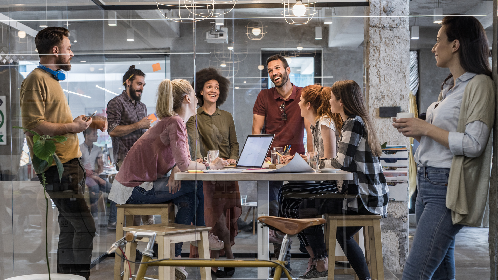 People in a modern office, collaborating around a table. Some smiling, using laptops, others standing nearby.