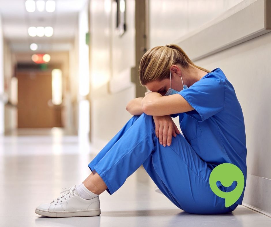 Nurse in blue scrubs, head in hands, sitting on floor in hospital hallway.