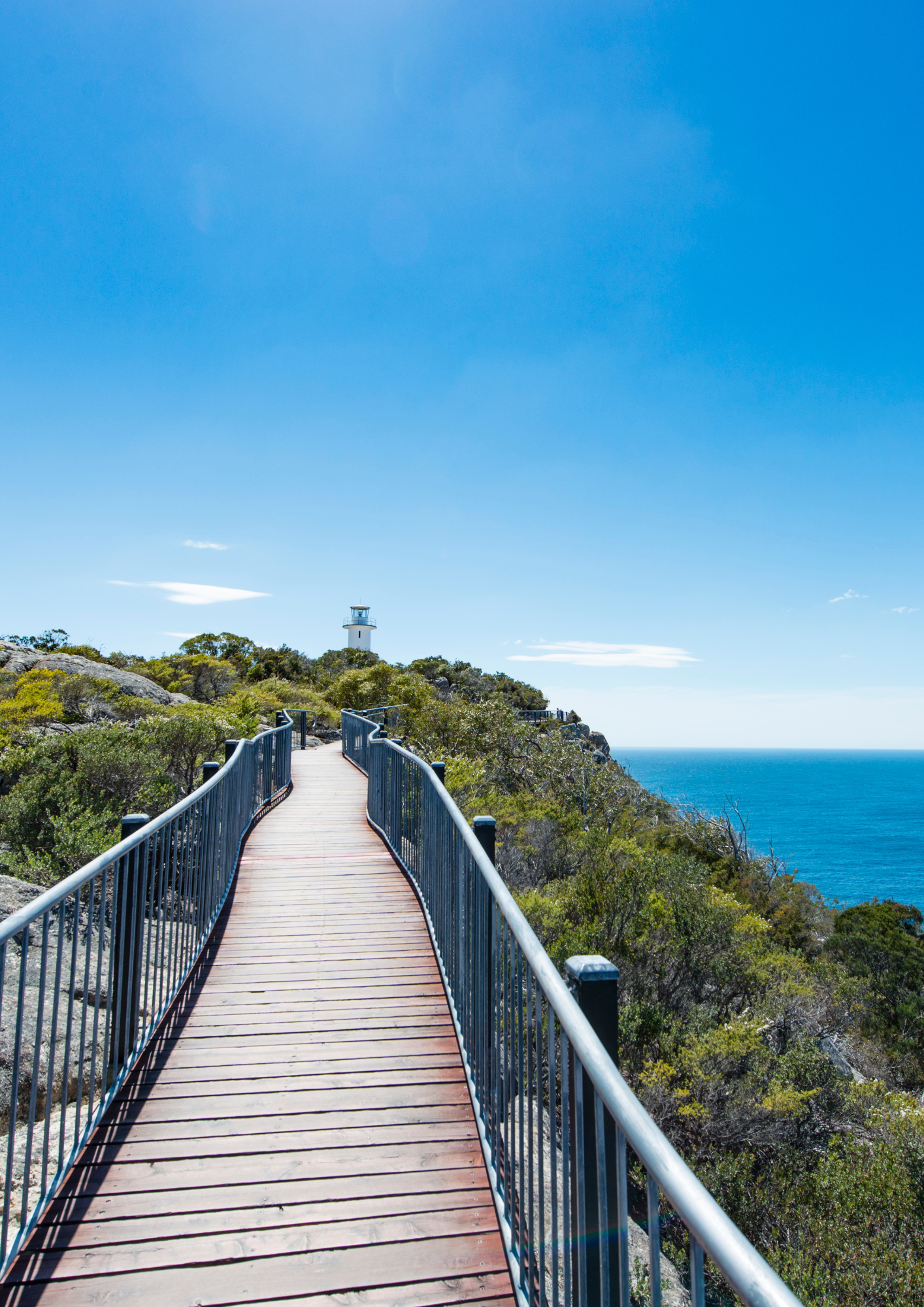 Wooden walkway leads to a lighthouse on a cliff, overlooking the blue ocean under a bright sky.
