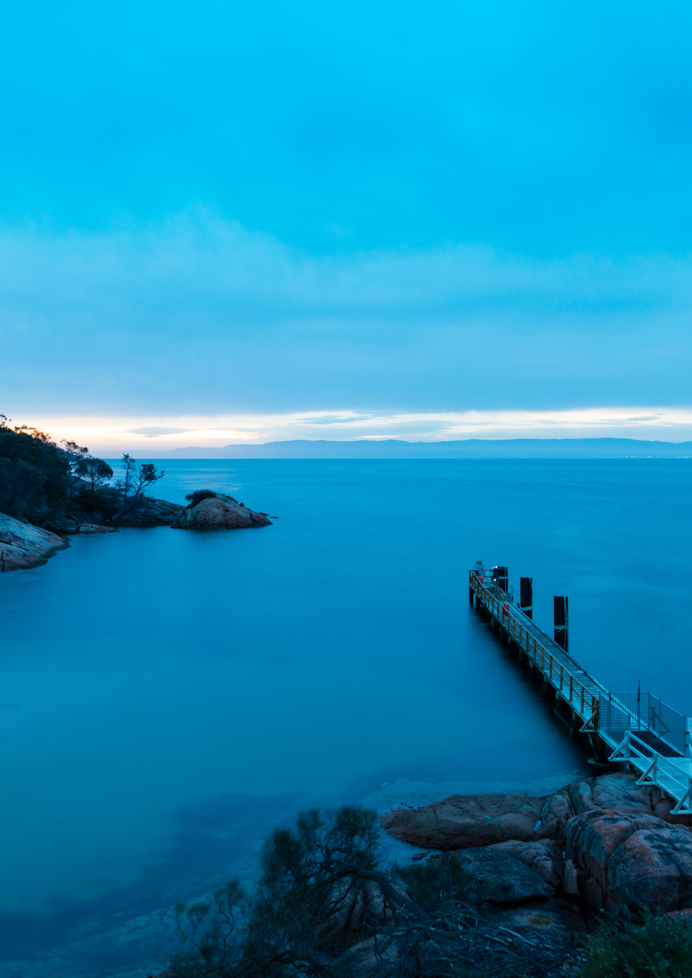 Wooden pier extends into a calm blue sea; rocky shoreline under a blue sky. Tasmanian landscape