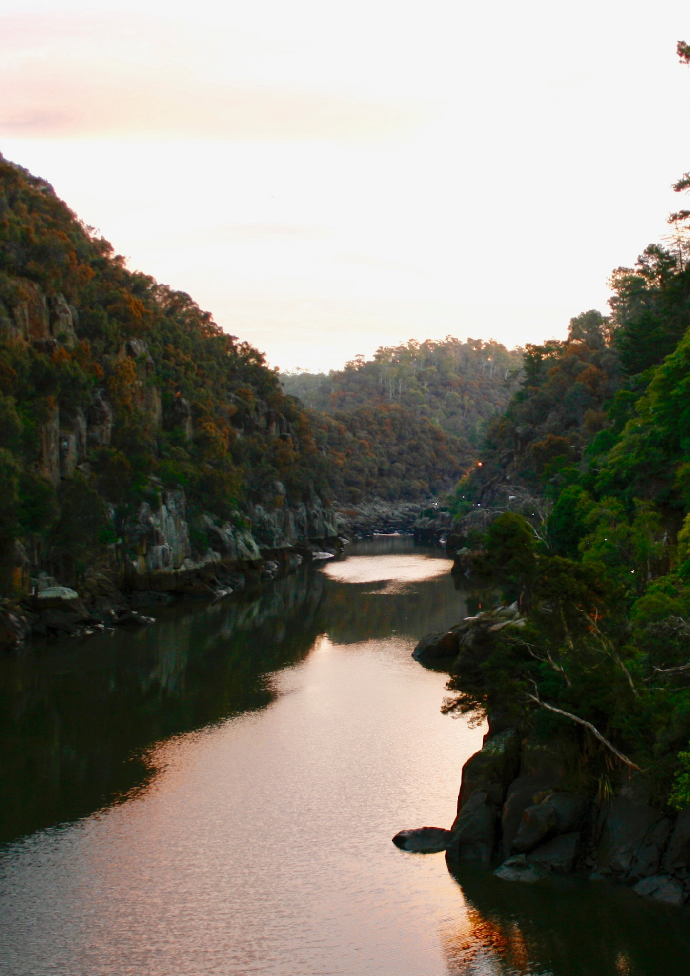 River winds through rocky gorge, reflecting golden light. Cliffs flank the water. Tasmanian landscape