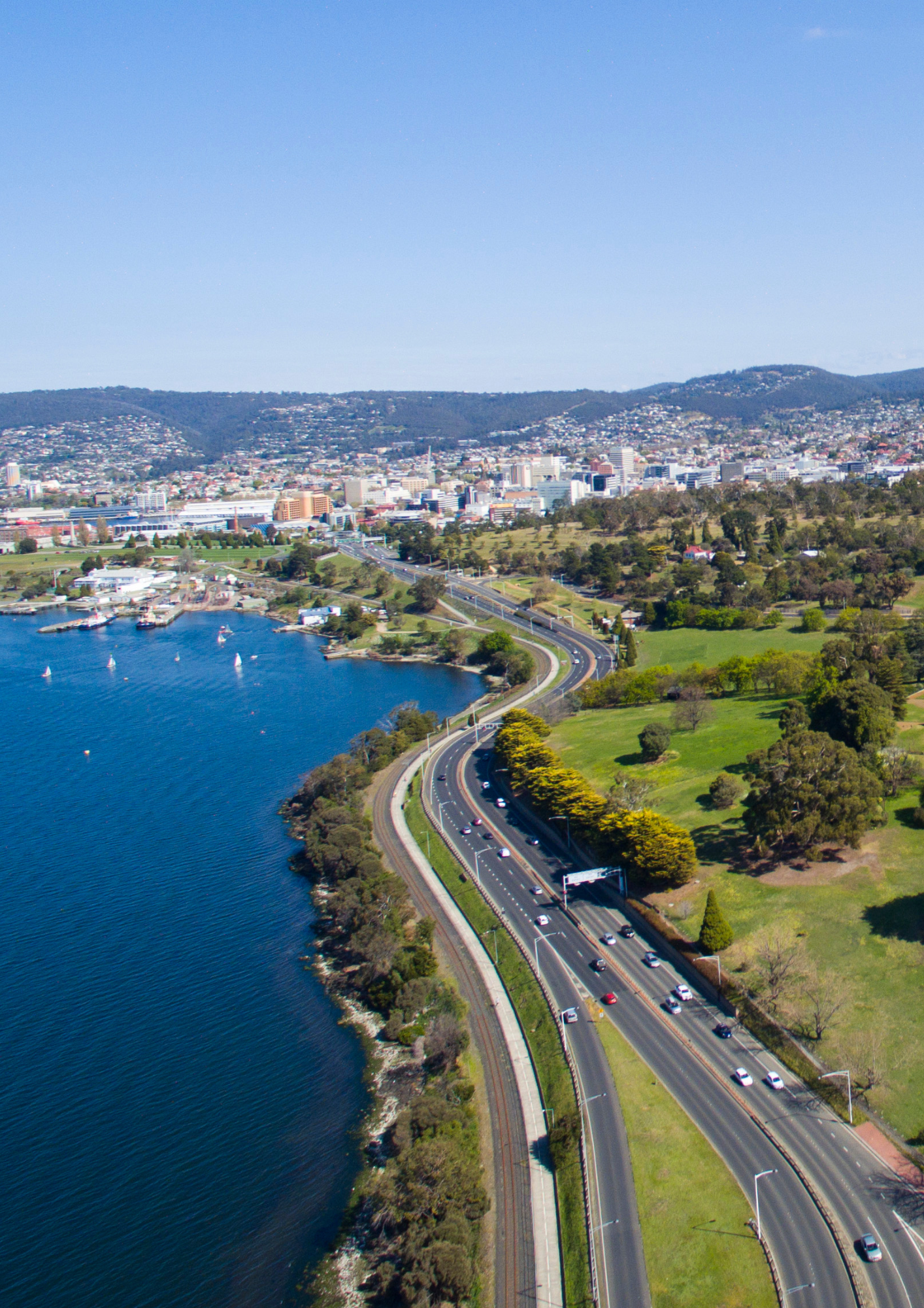 Road curves along a waterfront with city skyline in the distance on a sunny day. Tasmanian Landscape