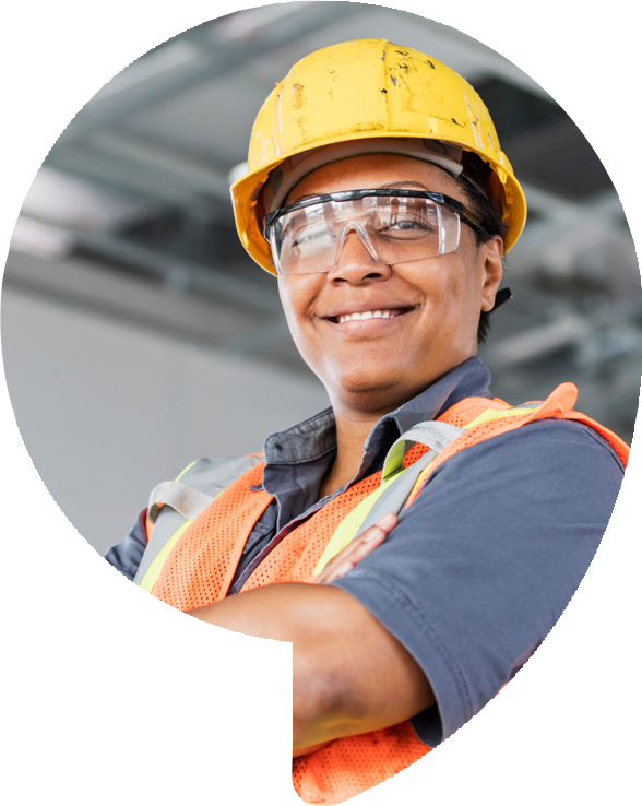 Construction worker smiles, wearing yellow hard hat, safety glasses, and orange vest, arms crossed.
