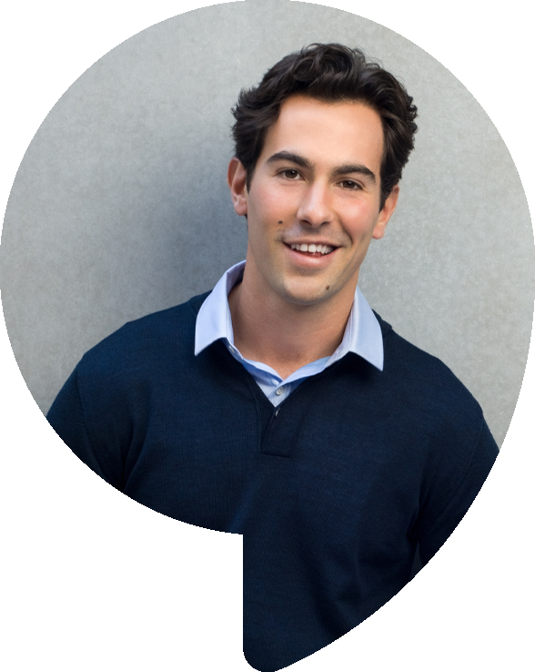 Smiling person in a blue sweater and collared shirt stands in front of a gray wall.