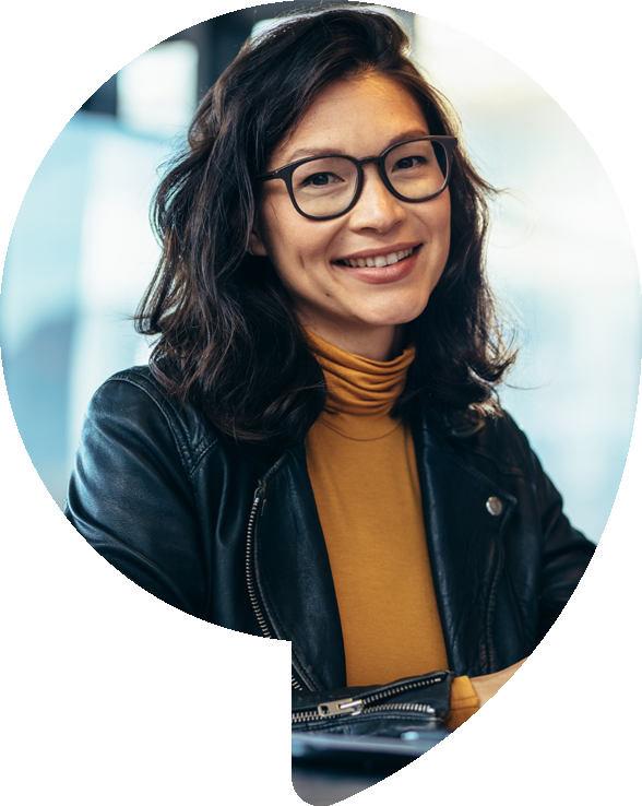 Woman with glasses, smiling, wearing a leather jacket and mustard-colored turtleneck, inside a building.