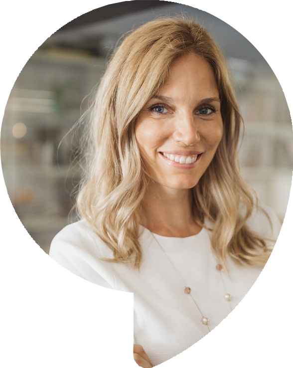 Blond woman smiles, wearing white top and necklace. Blurred office background.