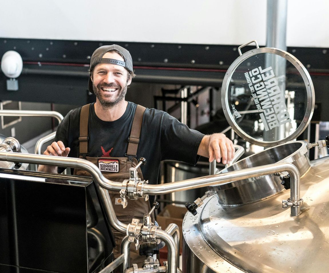 Man smiling next to large stainless steel brewing equipment in a brewery. He wears a hat and overalls.