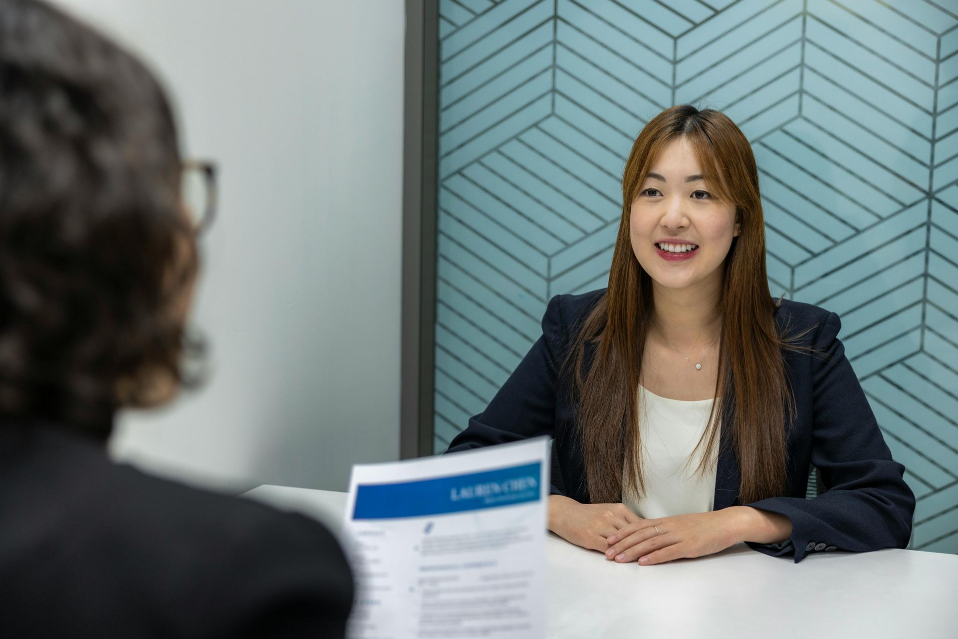 Woman in a blazer smiles during an interview. Resume on the table, blue and white geometric wall in background.