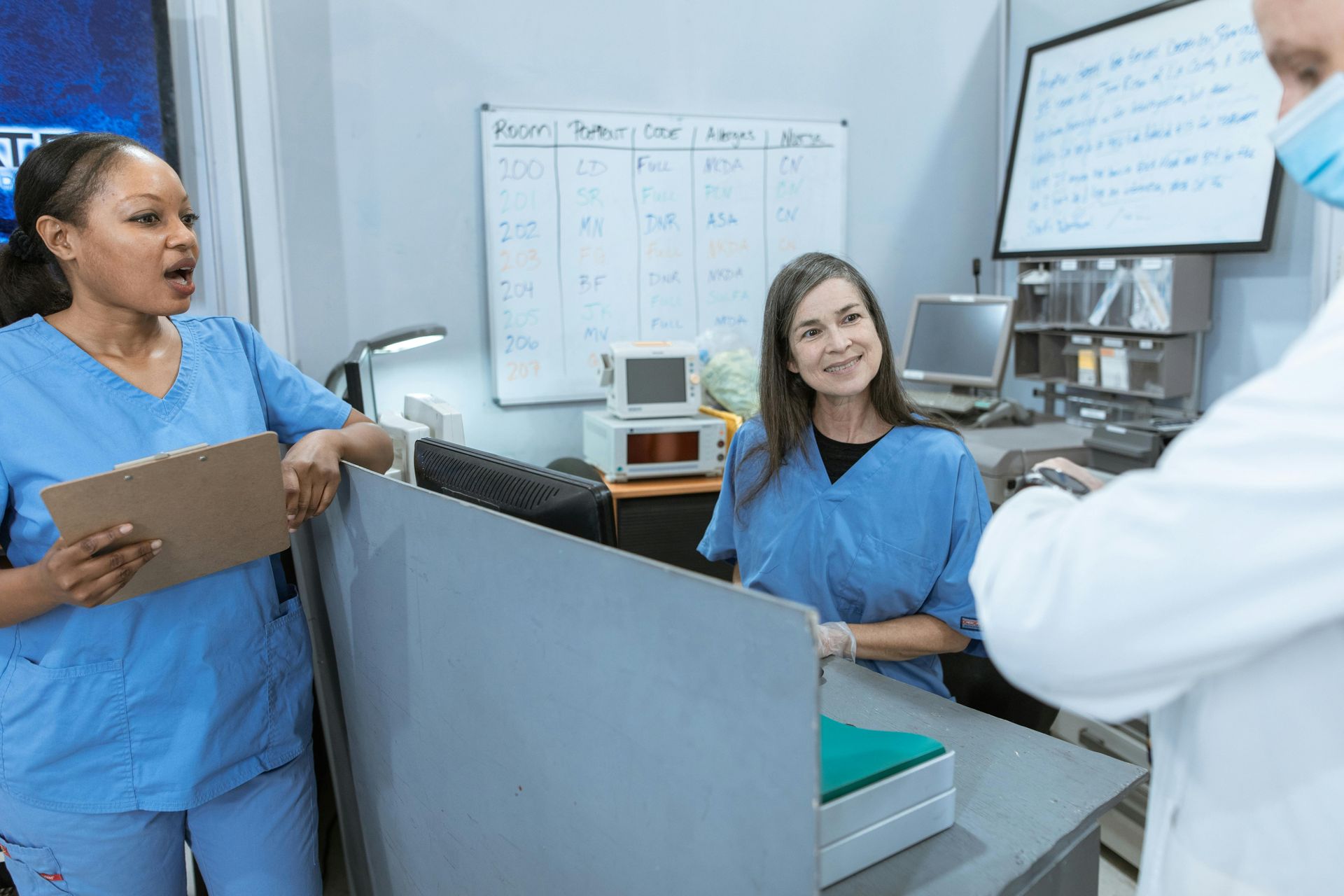 Medical staff in blue scrubs converse near a counter. A whiteboard and equipment are visible in the background.